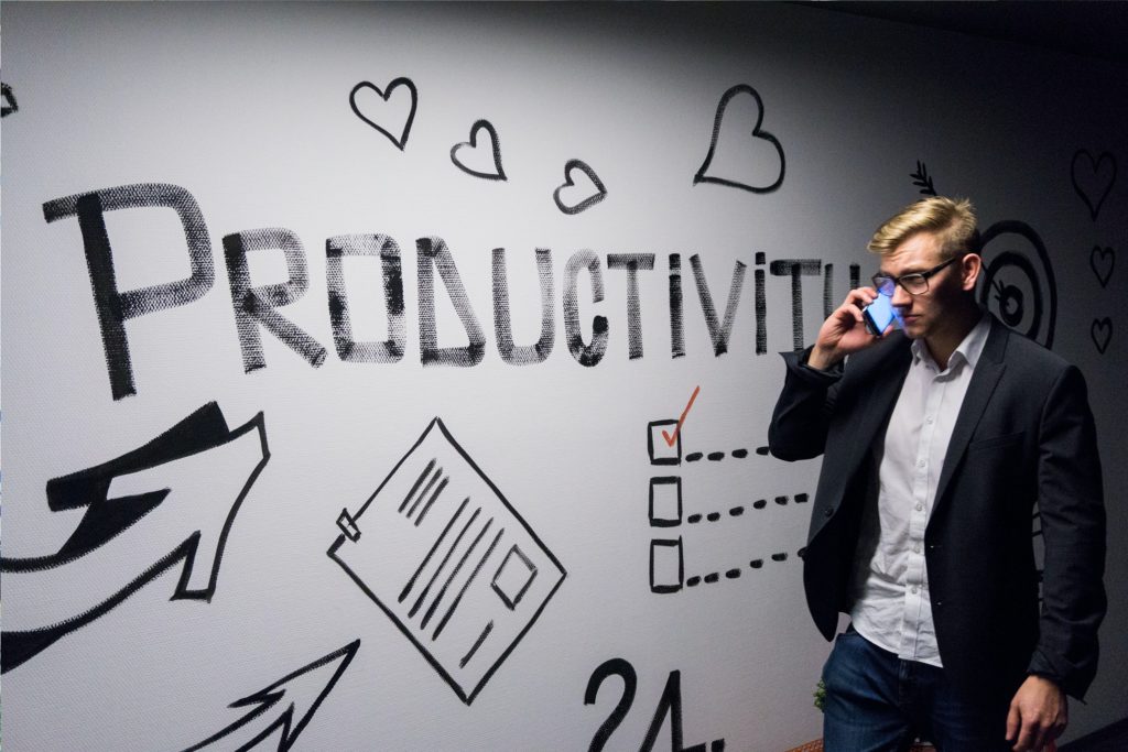 A man in a black jacket and blue jeans making a call in front of artwork that says productivity
