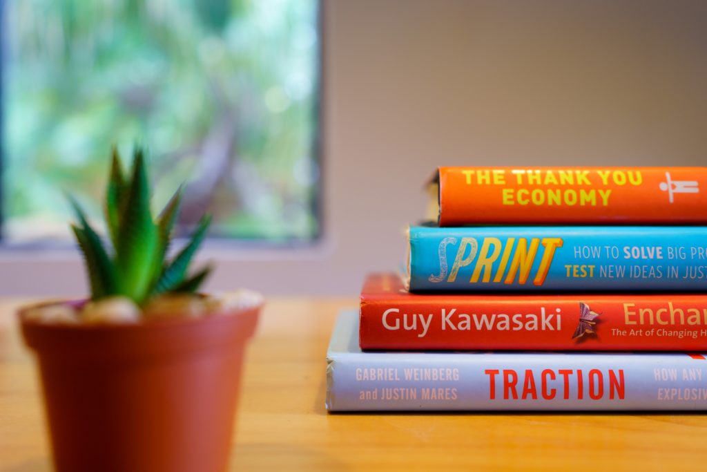 Four project management books placed on top of each other on a desk with a small potted cactus