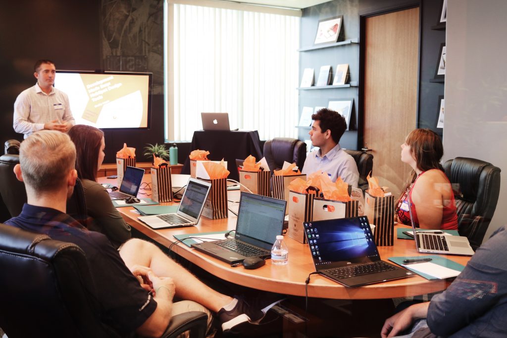 Six people at a meeting in a conference room with six laptops listening to a man give a presentation