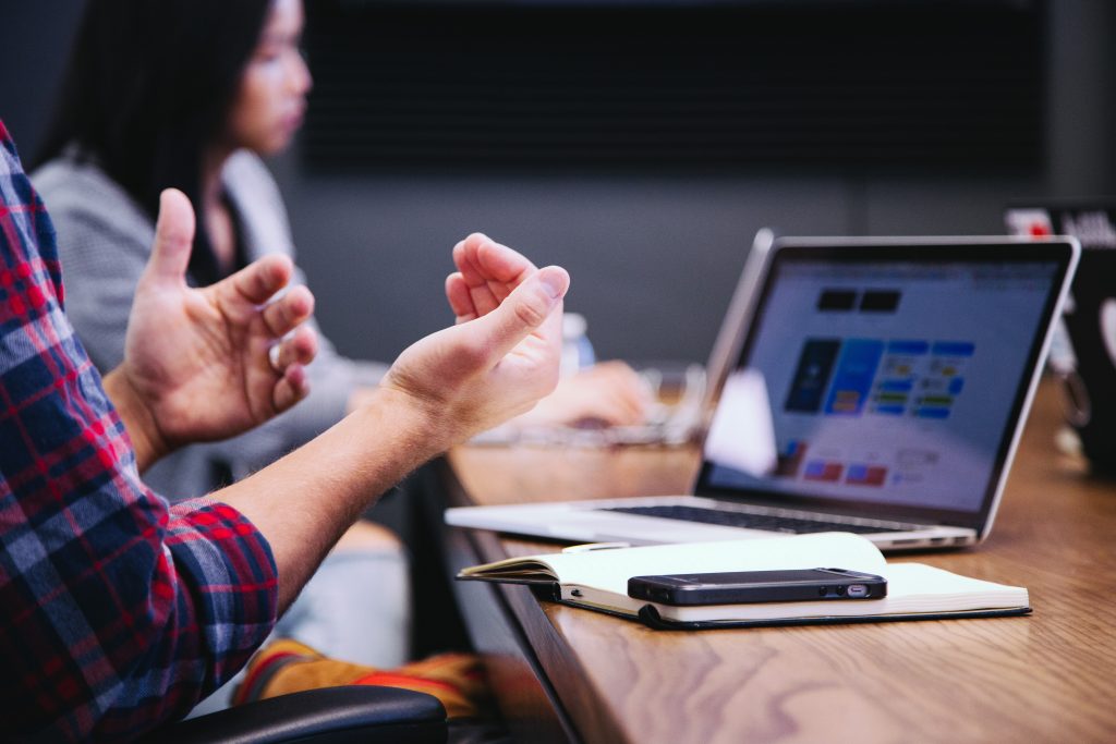 A pair of hands gesticulating in front of an open silver laptop on a wooden desk with a black phone on a small notebook