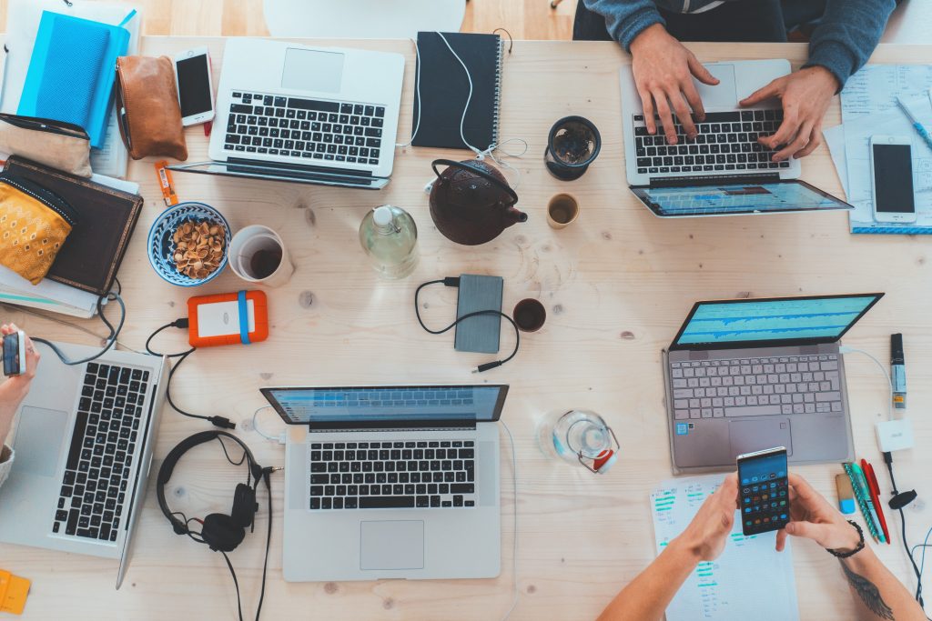 Five open silver laptops on a wooden table with three pairs of handing working from top view