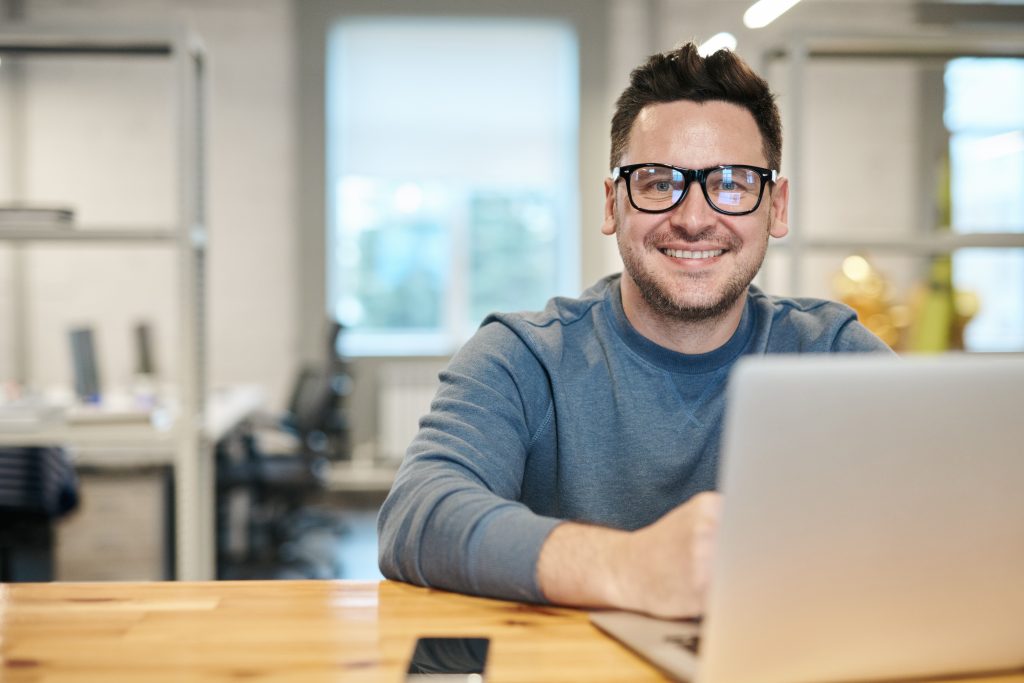 Man wearing eyeglasses sitting in front of a silver laptop in an open plan office