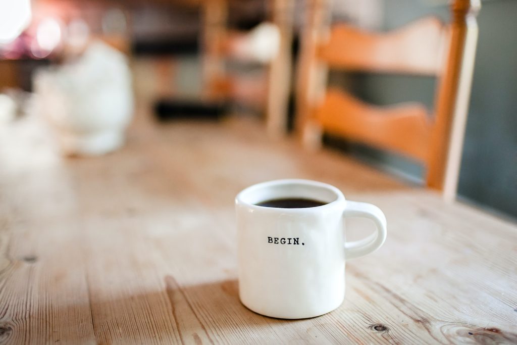White coffee cup filled with coffee with 'begin.' printed on it in caps in black, placed on a wooden table with two wooden chairs in background